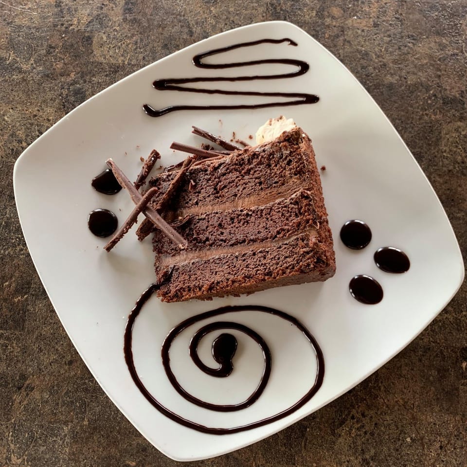 A slice of chocolate cake with chocolate shavings and syrup drizzle on a white plate.